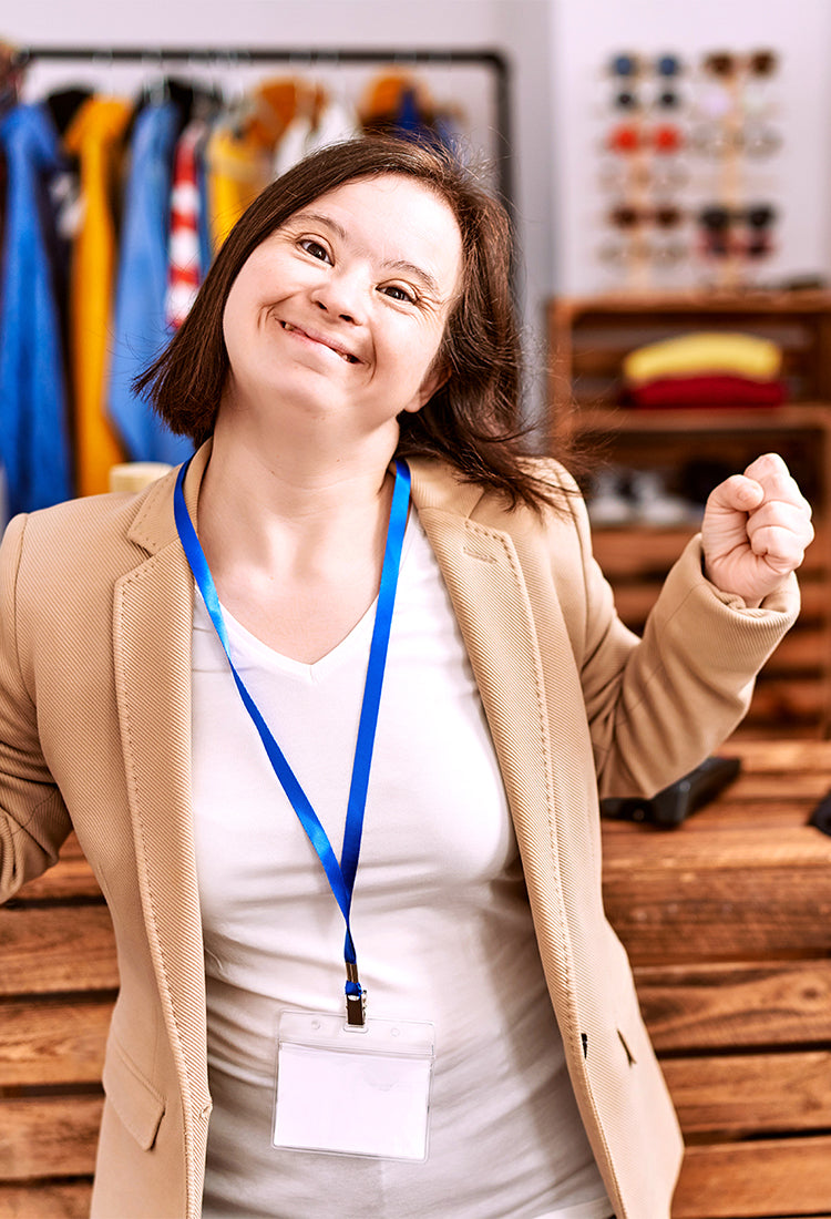 A young woman with Down syndrome is smiling brightly and raising her fists in a celebratory gesture, as if excited or triumphant. She is wearing a beige blazer over a white shirt with a blue lanyard around her neck. Behind her, various clothing items are displayed on racks, and shelves hold neatly folded clothes, suggesting she is in a retail or boutique setting. In the foreground, a computer monitor is visible on the right, indicating she may be working at the store or assisting customers.